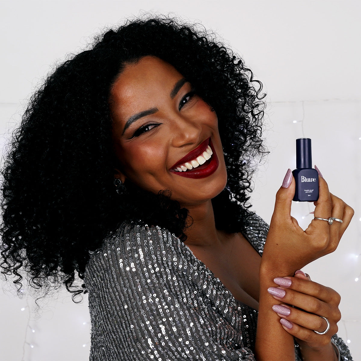 Modelo negra sorrindo em fundo branco segurando o esmalte Dreams do kit celebrate e com o esmalte aplicado nas unhas. 